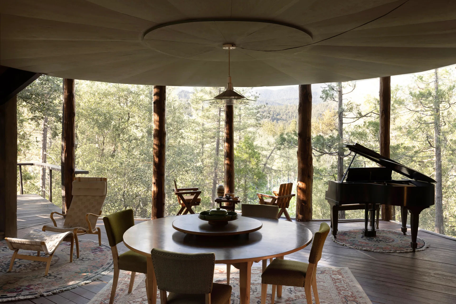 Circular main room with radiating wood-paneled ceiling and glass pendant over a round dining table, chartreuse chairs on a patterned rug, natural log columns, black grand piano, lounge chair with ottoman, and open views to pines and distant hills