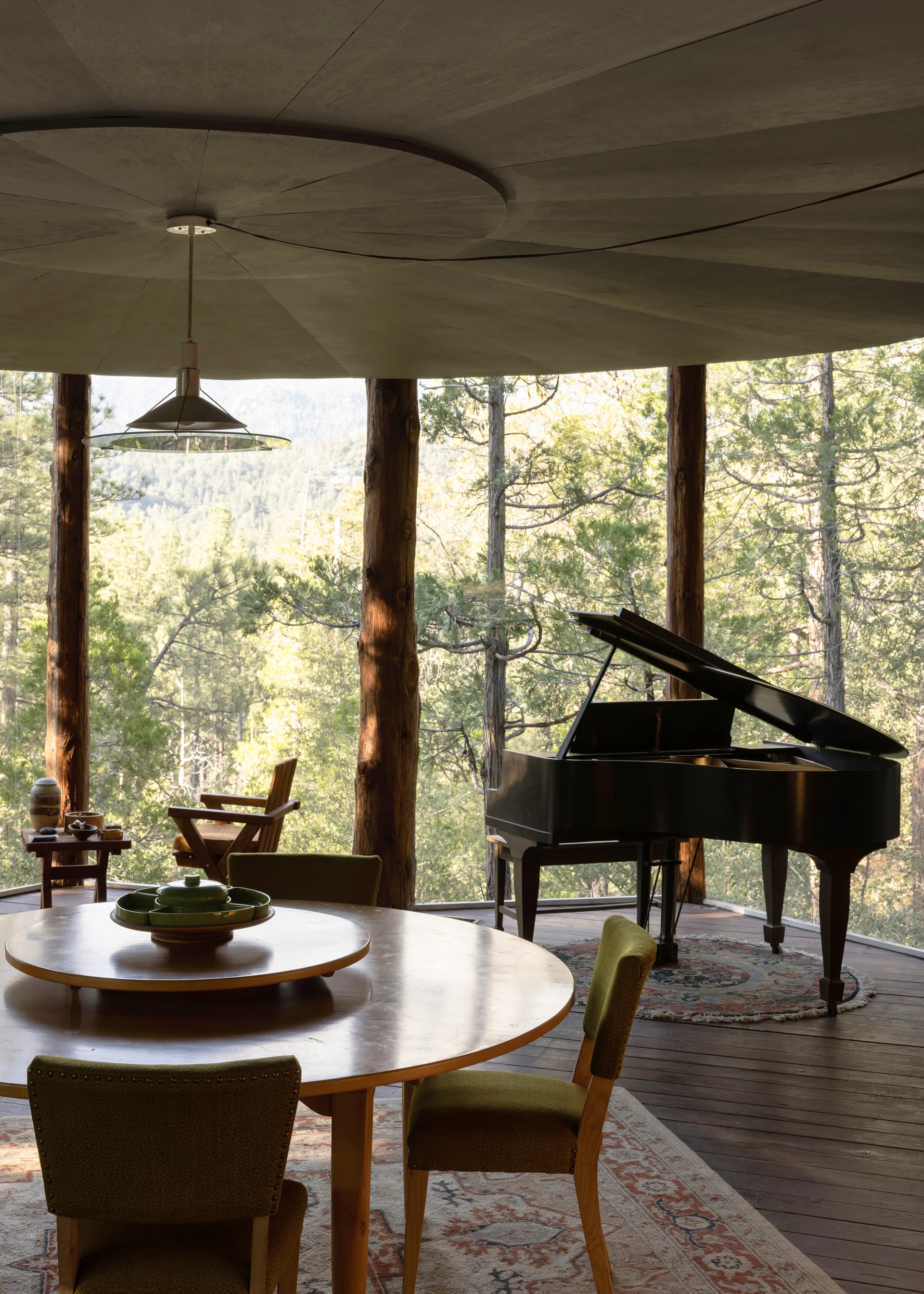 Dining area: round wood table with lazy Susan and green ceramic serveware, chartreuse upholstered chairs, starburst ceiling panel and cone pendant, grand piano beyond the log columns, evergreen forest and mountain through the glass