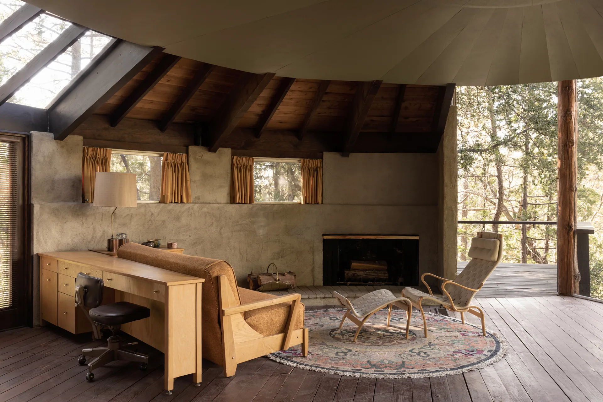 Living and work space with textured plaster walls, dark beamed ceiling and skylight, long wood desk with leather chair and tall lamp, bentwood lounge and ottoman on a patterned rug, fireplace with stacked wood, mustard curtains on small windows, and an opening to a wood deck among trees