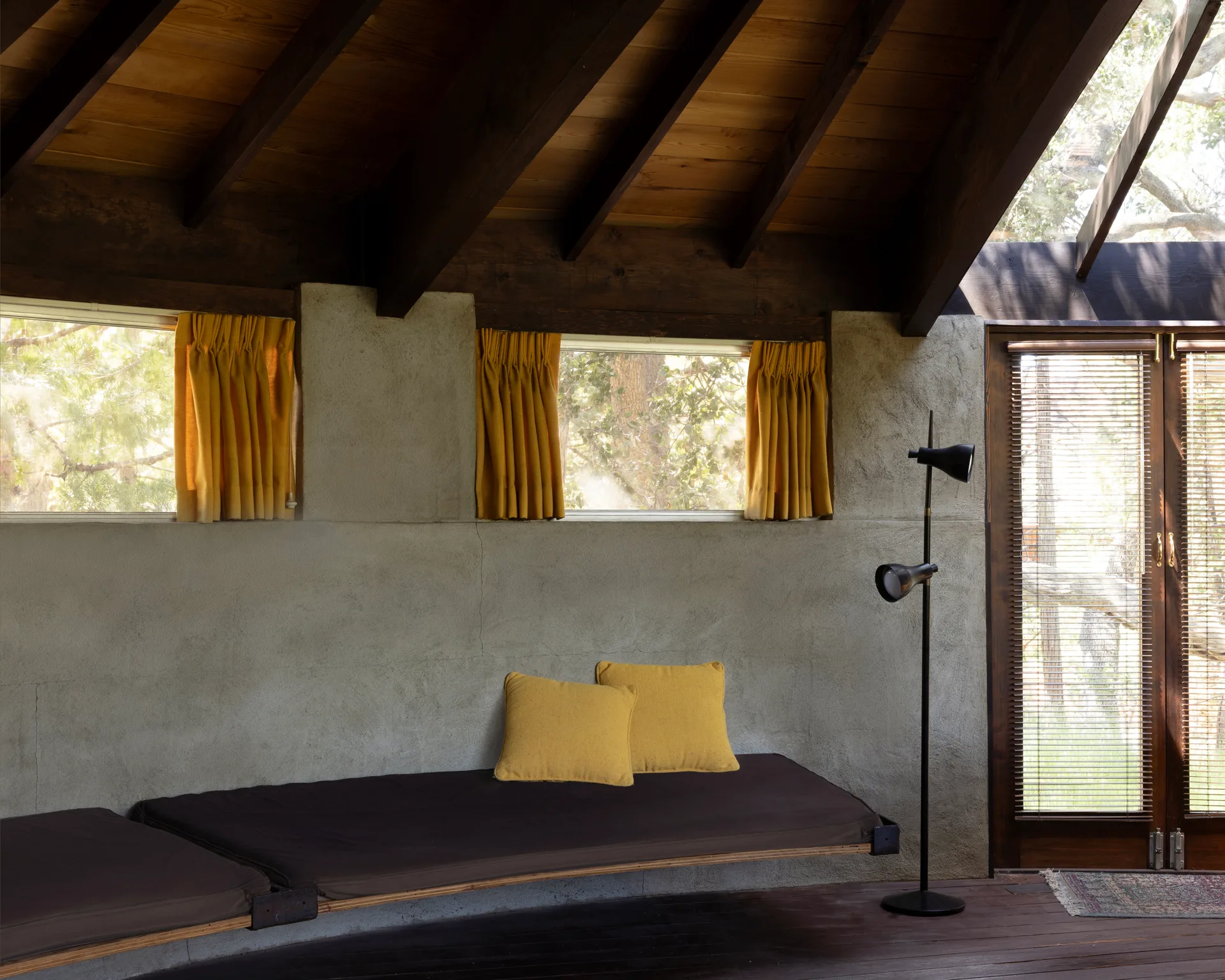 Built-in cantilevered benches along a textured gray wall with cushions and mustard throw pillows, two small horizontal windows with matching curtains, black dual-head floor lamp, dark wood flooring, and a glass door opening to green trees