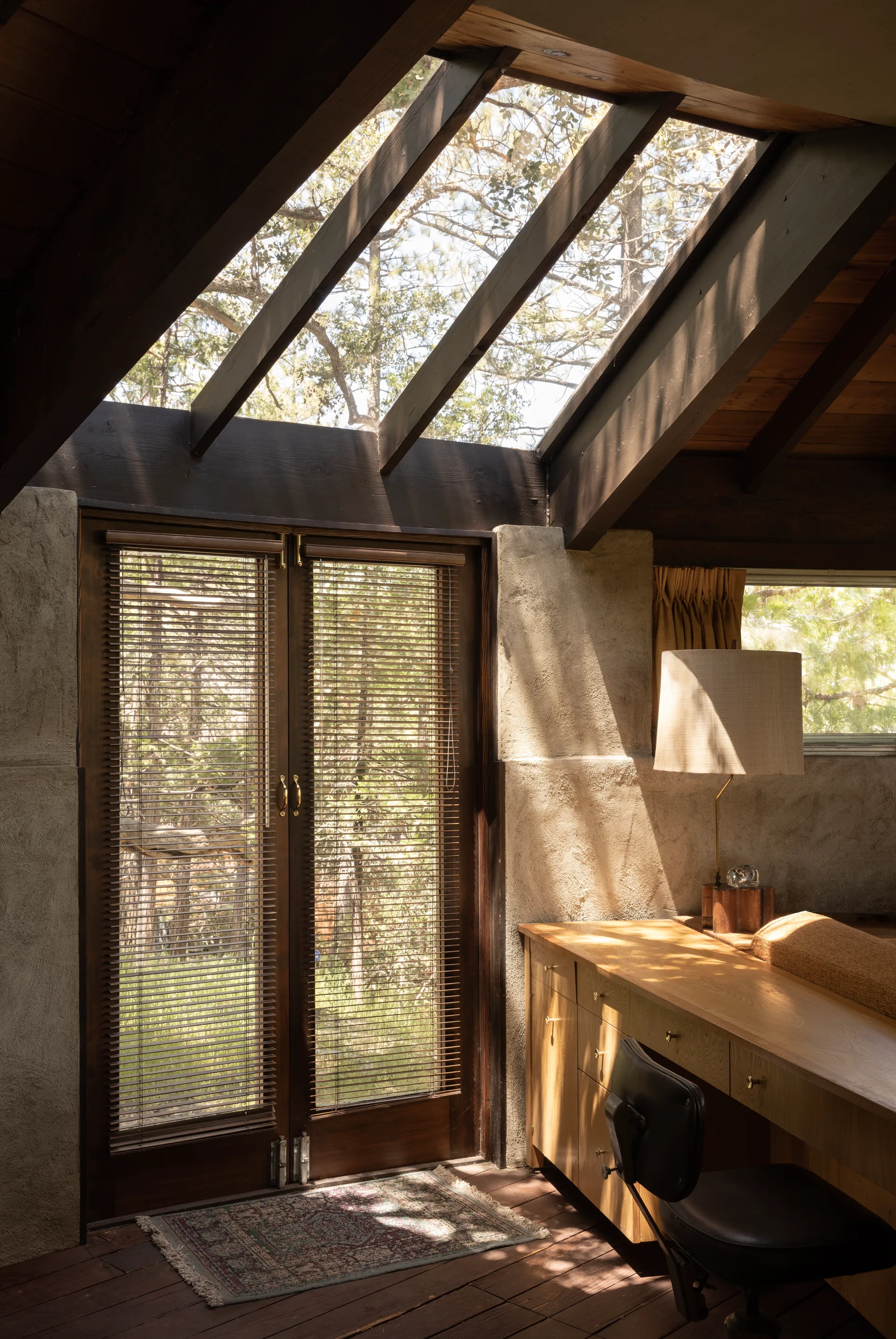 Office corner with sloped wood ceiling, large skylight showing branches, pair of wood French doors with blinds on a patterned rug, long dresser-style desk with cylindrical shade lamp and leather desk chair