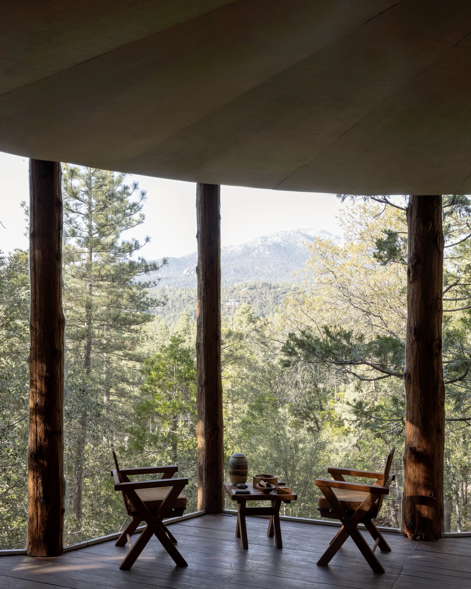 Glass corner between log columns with two wood folding chairs and a small side table holding ceramics, dark plank floor and sloped ceiling, looking out over evergreen forest to a rocky mountain peak under pale sky