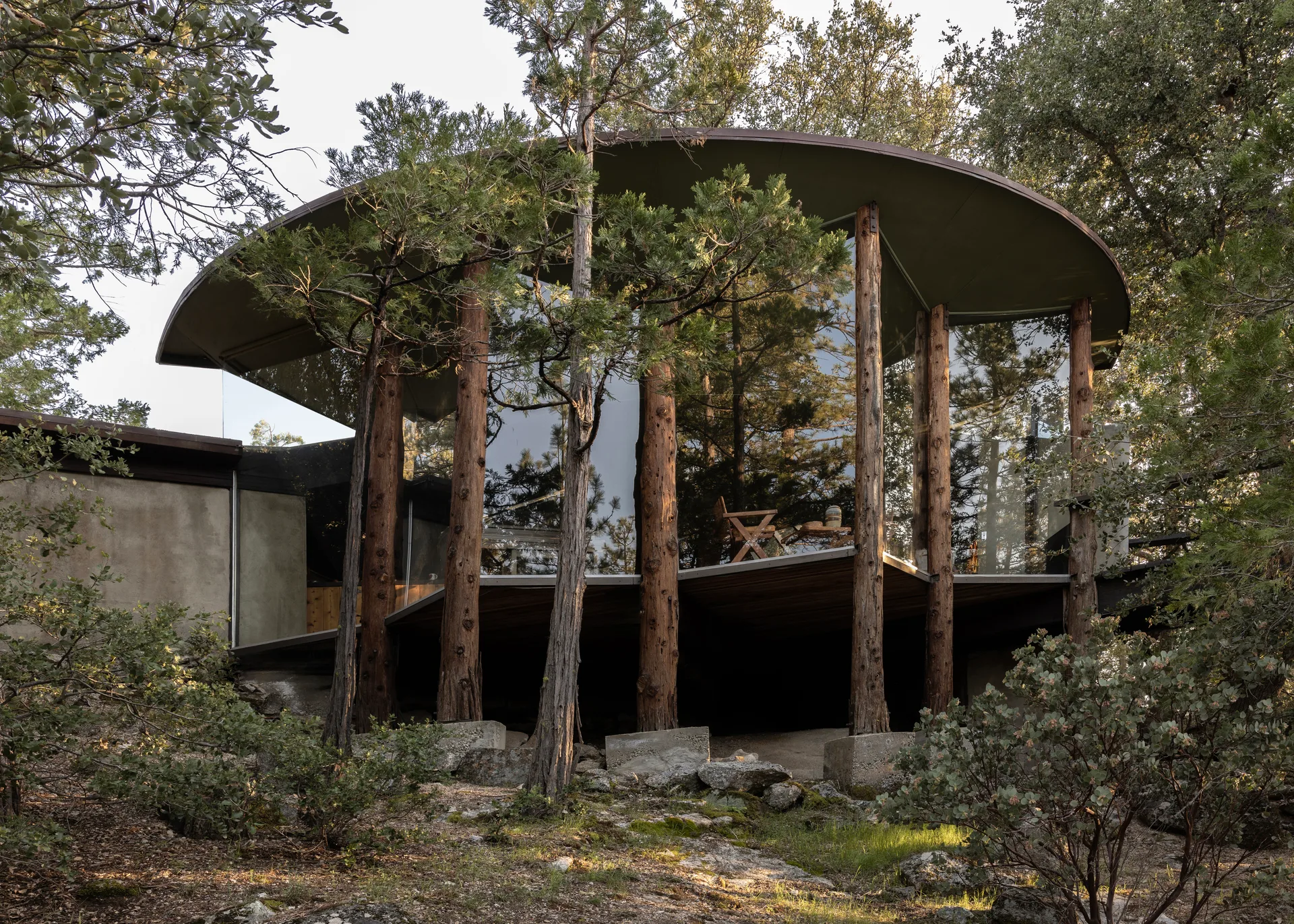 Wide landscape view of the glass drum and flat circular roof on log columns, stucco wing at left, rocky leaf-strewn slope and pines; folding chair and table glimpsed through the glass (7×5 aspect crop of one photograph)
