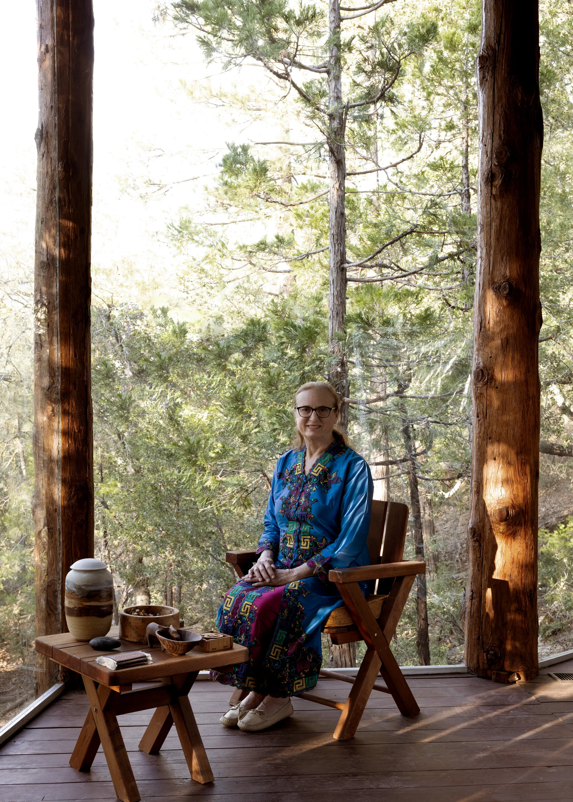 Nancy Pearlman seated on a wood armchair on the cabin deck between two log columns, wearing a bright blue caftan with colorful embroidered bands; side table with lidded ceramic jar and bowls, sunlit pines behind