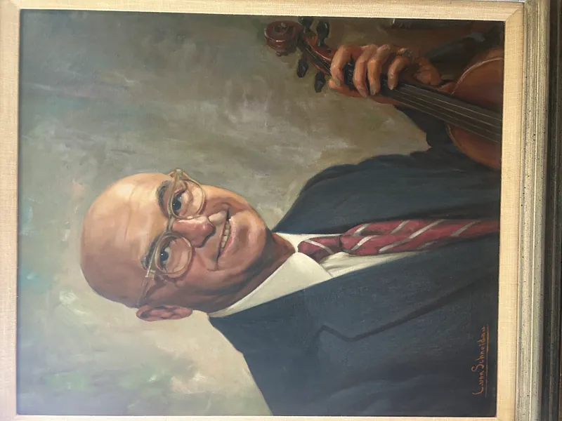 Portrait of Dr. Carl Kenneth Pearlman, a smiling bald man wearing glasses, a dark suit, white shirt, and red and white striped tie, holding a violin