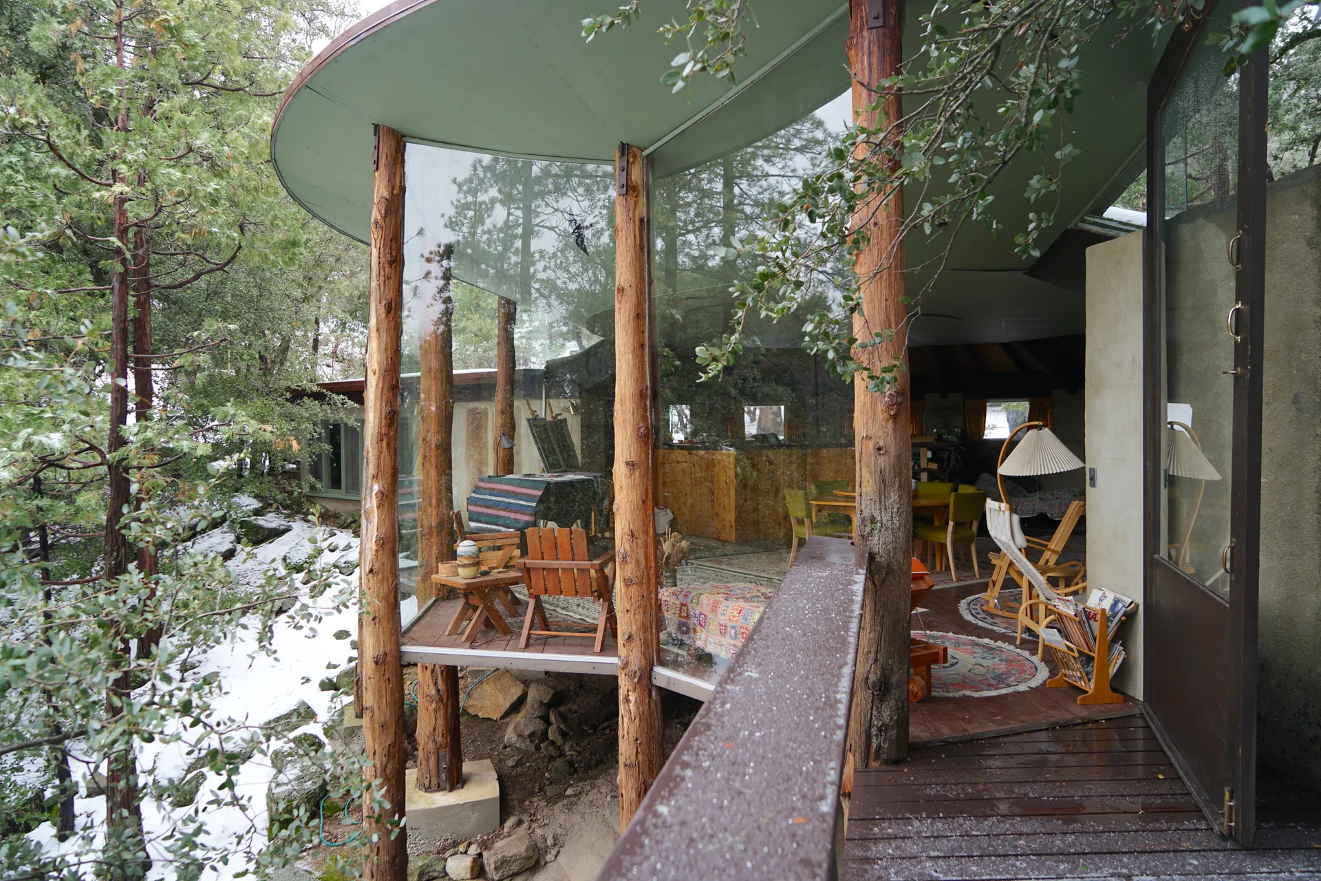 Snow-dusted deck and open door to the cabin, with interior seating, ceramic vessels on a wooden table, patterned rugs, and a pleated floor lamp visible through the curved glass