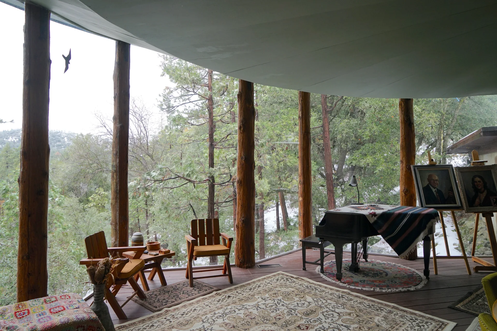 Interior with natural log pillars, slatted wooden chairs by the windows, grand piano draped with a striped throw, and two framed portraits on easels with forest view beyond