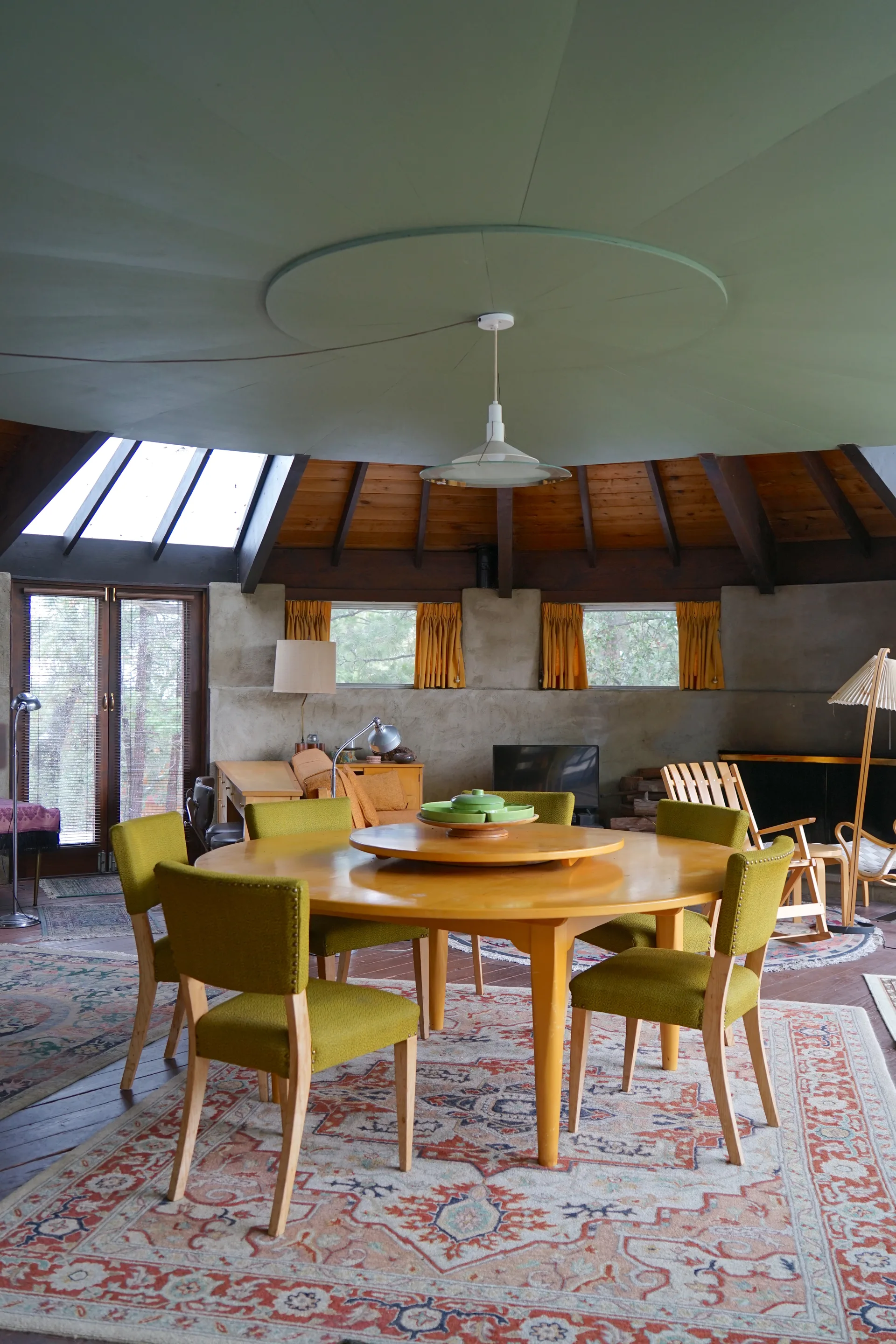 Dining area with round table and chartreuse chairs, lazy Susan with green ceramic dishes, sage green curved ceiling and pendant light, skylights and orange curtains, oriental rugs and rocking chairs