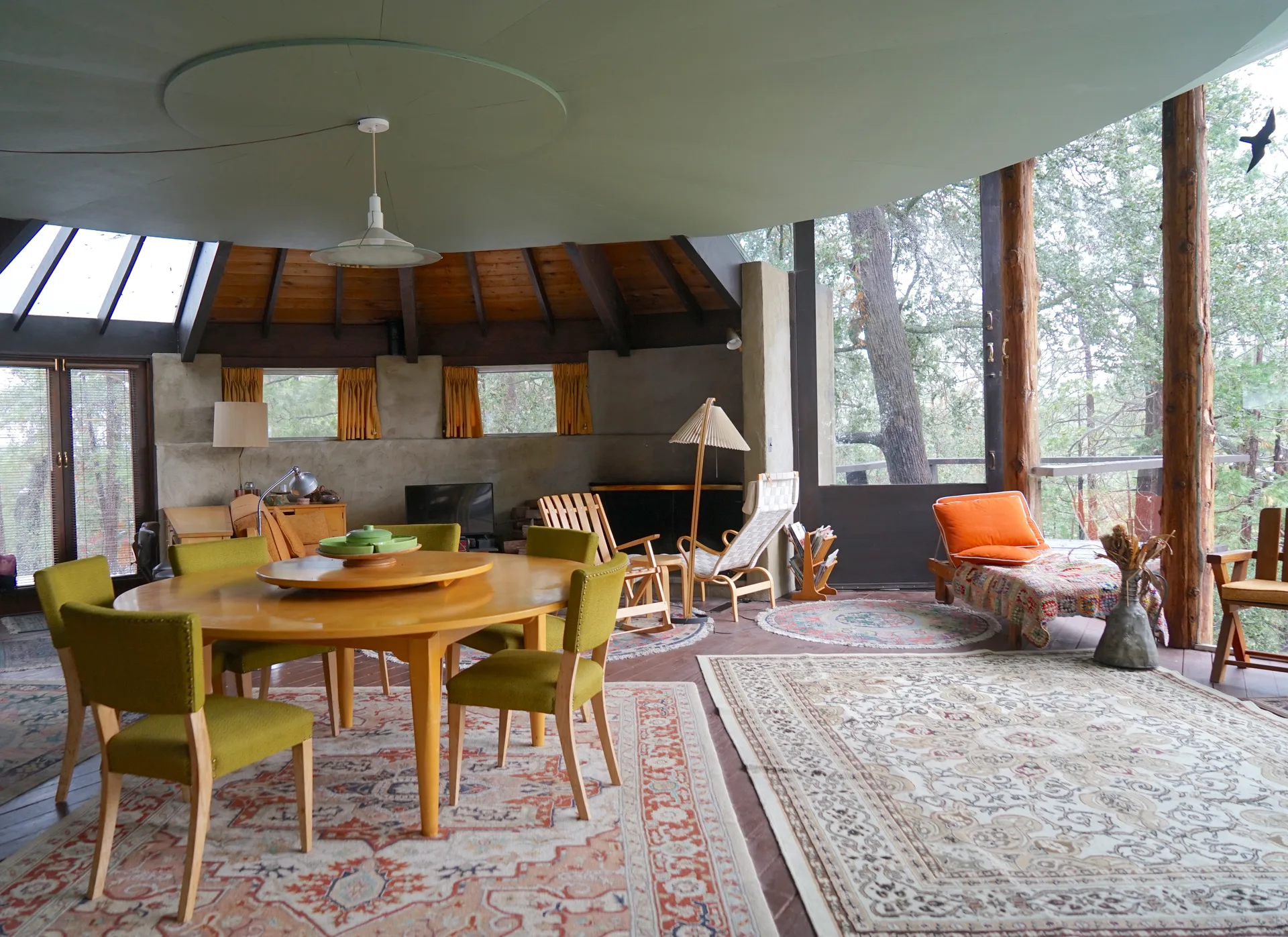 Circular room with oval dining table and chartreuse chairs, orange-gold curtains, chaise and wooden lounge seating, pleated floor lamp, and floor-to-ceiling windows with forest view
