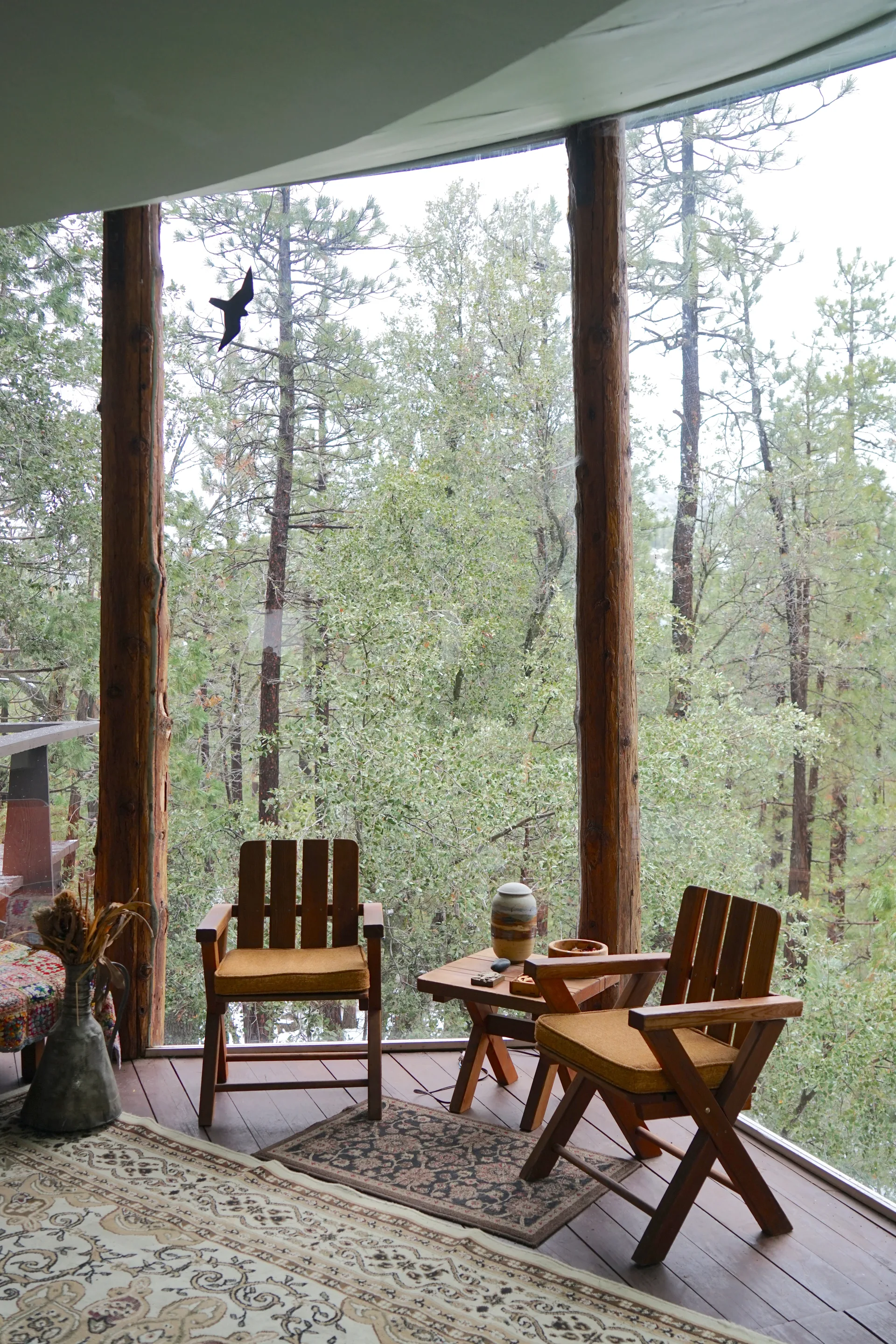 Seating by the windows: two wooden chairs with mustard cushions and a small table with ceramic vase, framed by log columns and light green ceiling, with forest view