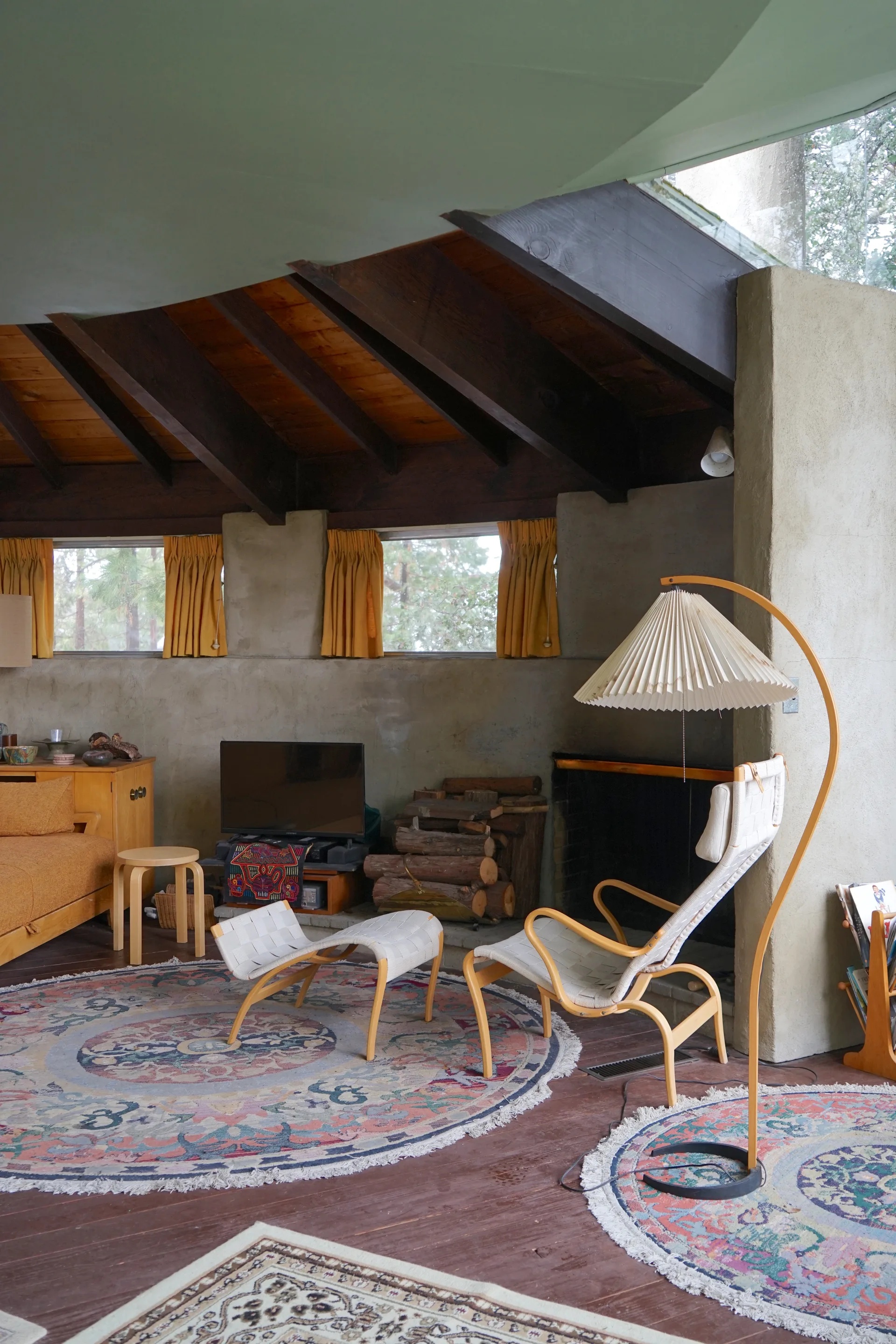 Living area with curved sage green ceiling and dark beams, mid-century curved lounge chairs on circular rugs, yellow curtains, fireplace with stacked wood, and pleated floor lamp
