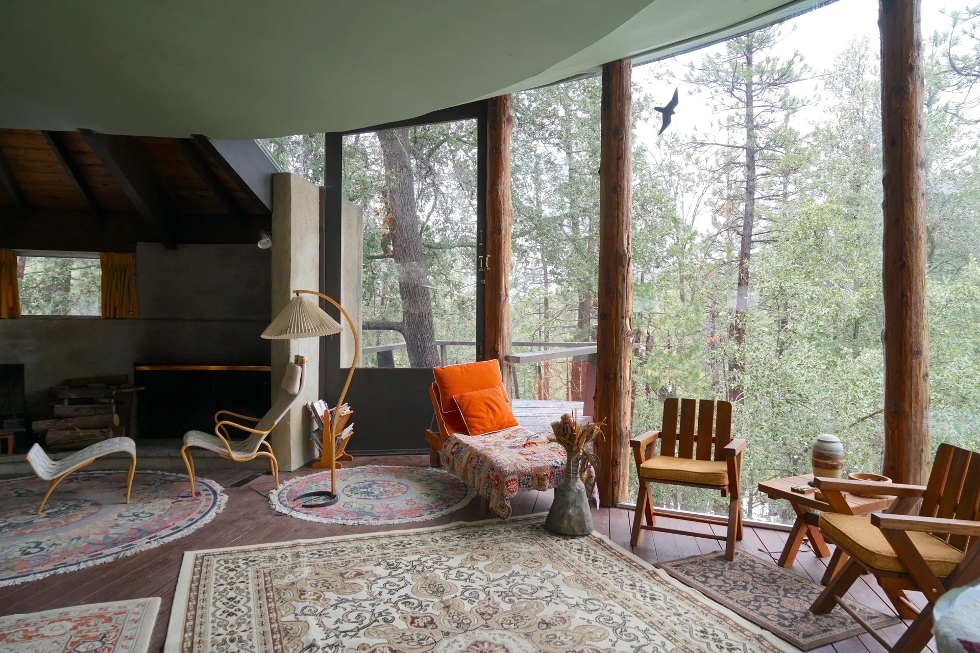 Wide interior view with curved floor-to-ceiling windows and forest vista, mix of modern and rustic seating—chaise, armchair, orange-cushioned lounge with throw, Adirondack chairs—ornate rugs, pleated floor lamp, and fireplace with stacked wood