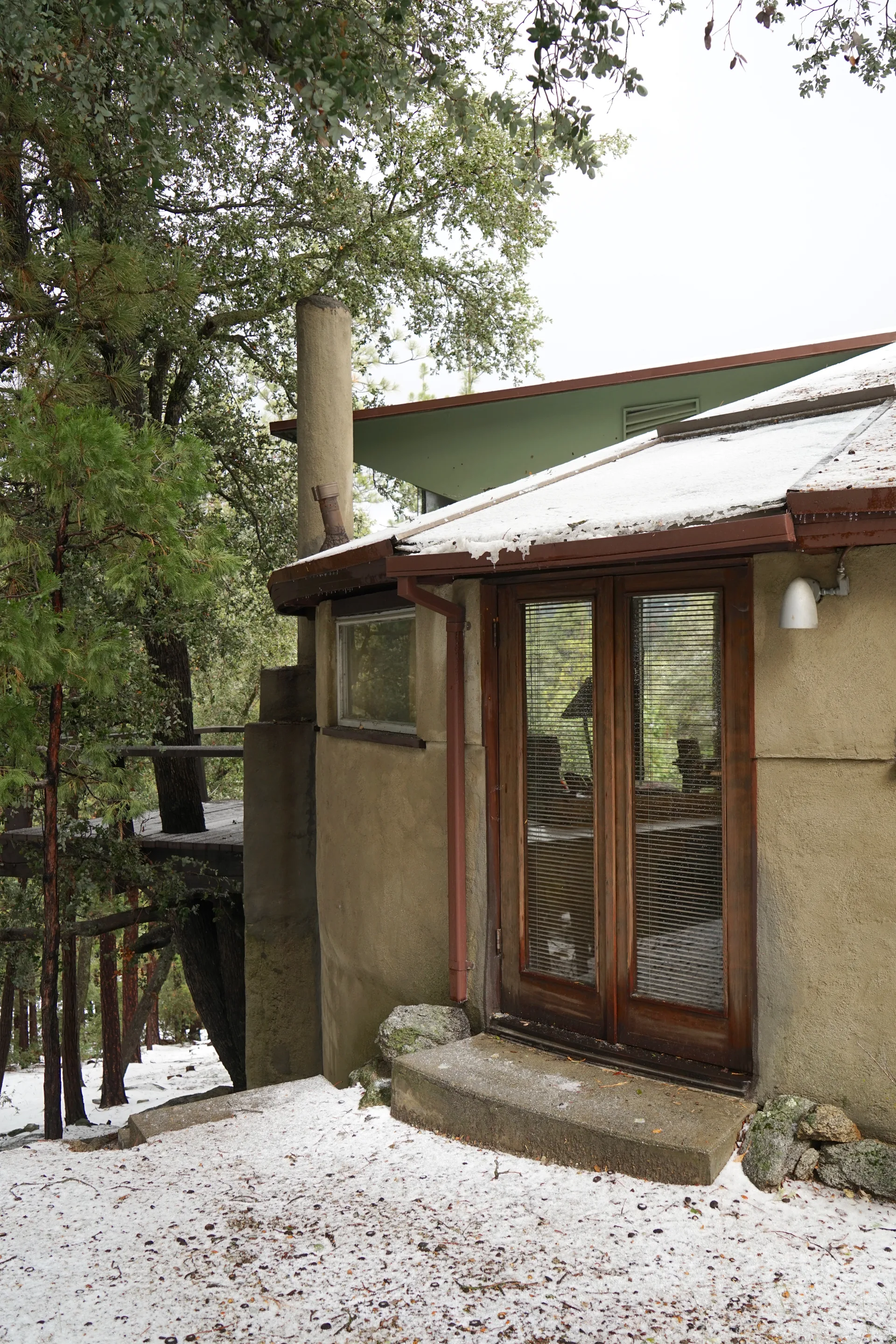 Winter view of the cabin’s side entrance: tan stucco walls, double wooden doors with glass panes and blinds, cylindrical chimney, and snow-dusted roof amid trees