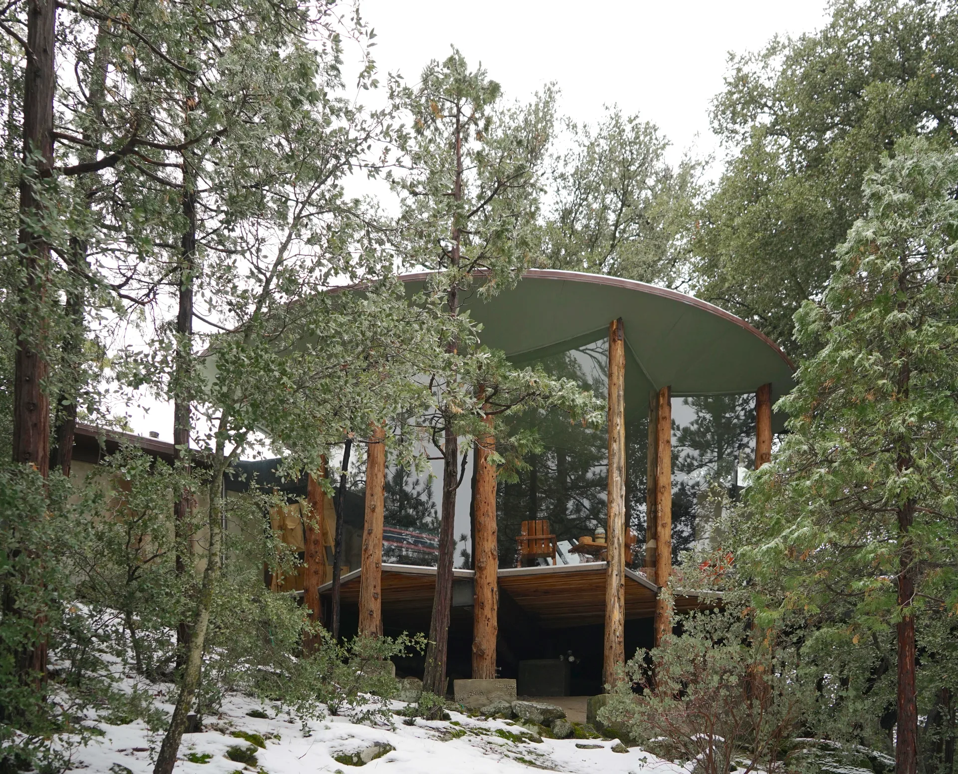 Exterior showing the curved olive-green roof and tree-trunk supports, floor-to-ceiling glass and deck with two Adirondack chairs and table, in snowy forest