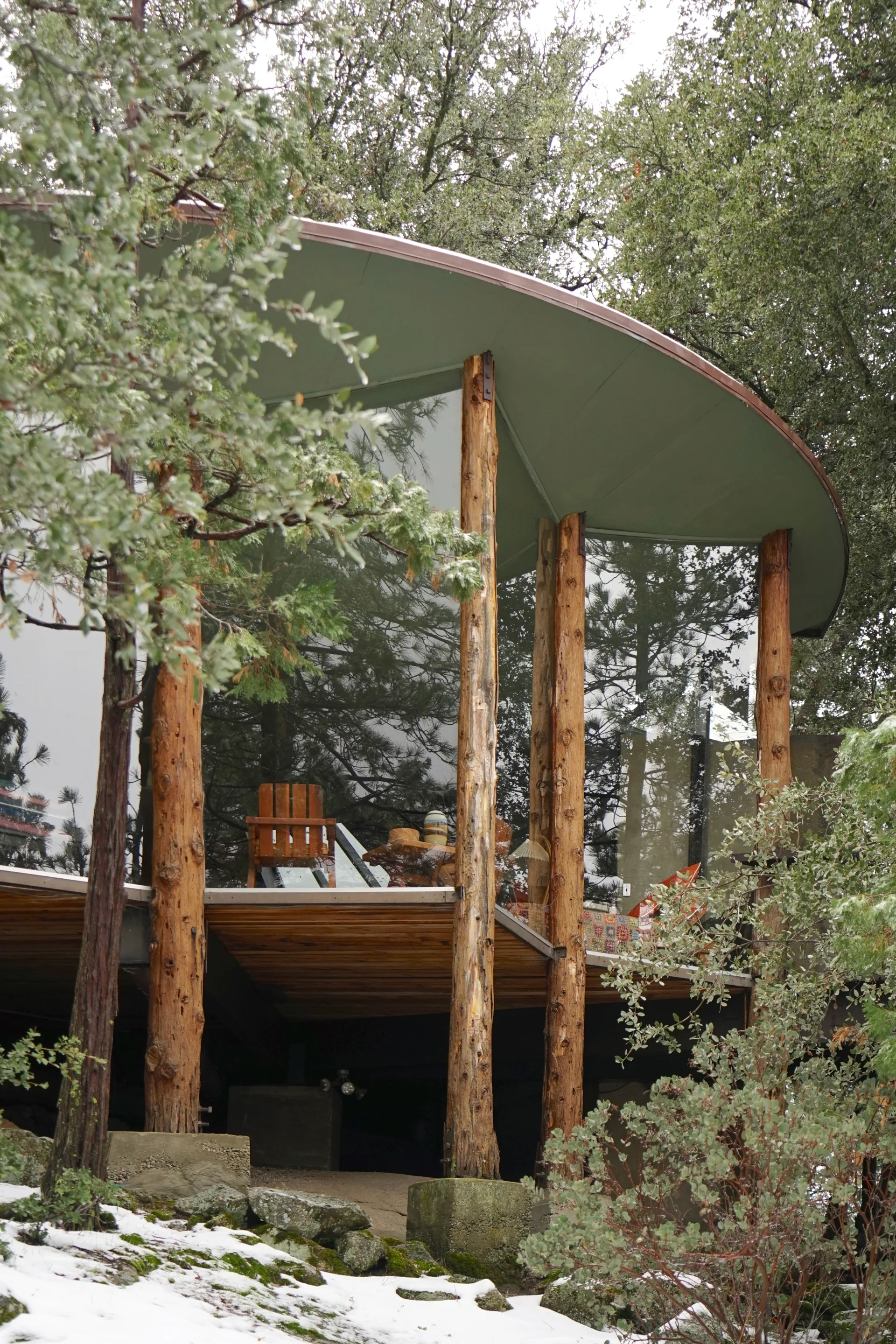 Curved sage green roof with dark trim, reflective glass walls and log columns, deck with Adirondack chair and pottery, snow and rocks in foreground