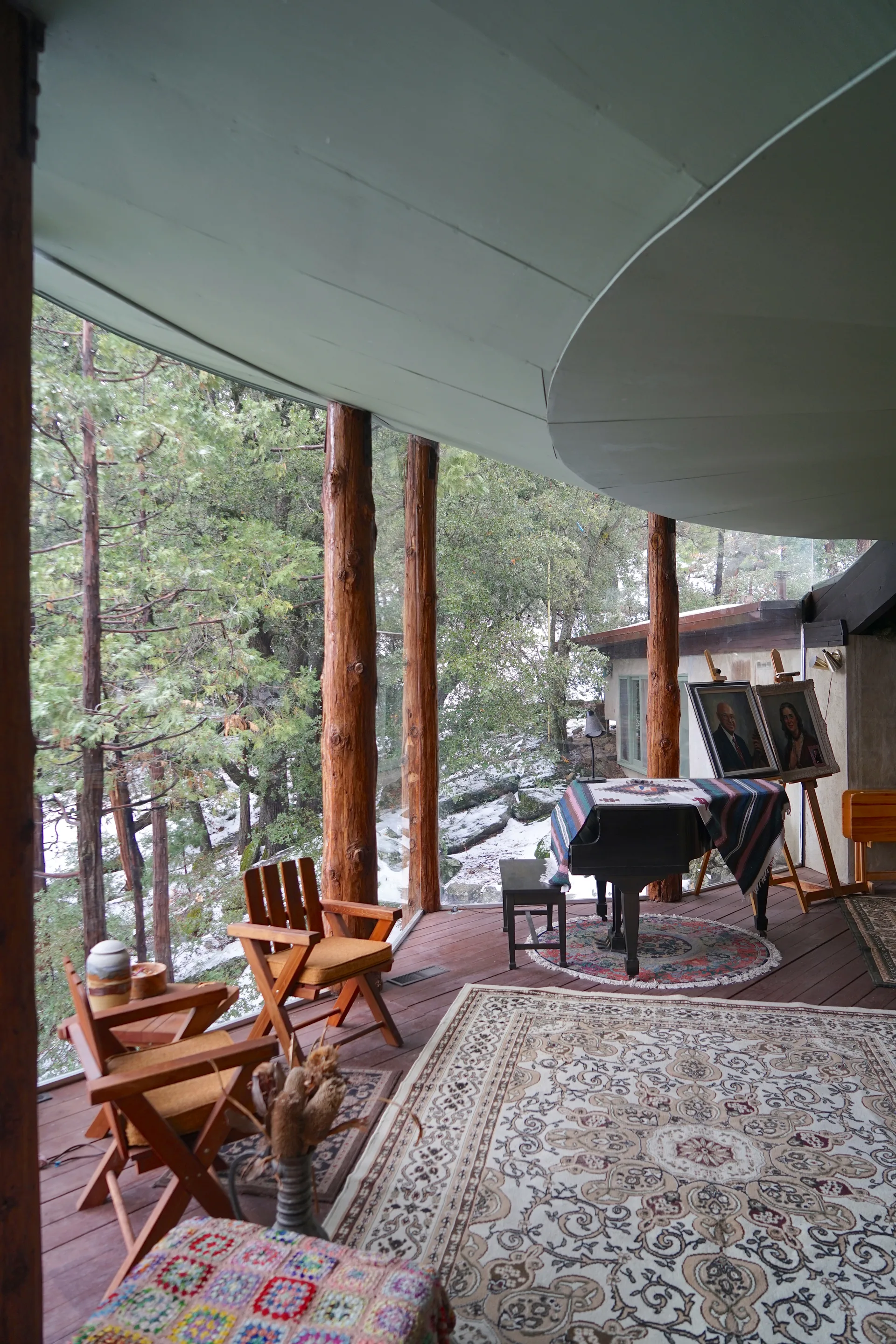 Furnished outdoor deck with rugs, wooden folding chairs, small upright piano covered with a striped serape, two easels holding framed portraits, pottery on a side table, and forest view in snow